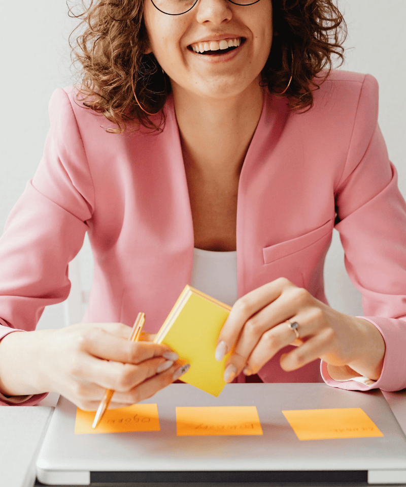 Brown haired woman, smiling, holding a stack of yellow post it notes above a folded laptop with three strategy notes already stuck to it.